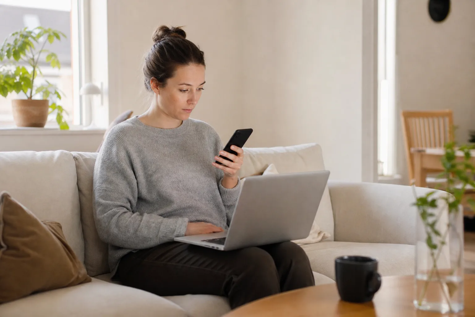 Person jämför billigaste mobilabonnemanget på laptop och mobil i soffan hemma
